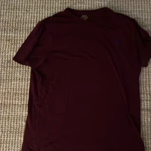 Vinröd t-shirt Polo Ralph Lauren XL - Vinröd t-shirt från Polo Ralph Lauren i storlek XL. Slim fit-modell med klassisk rund hals och blå broderad logga på bröstet. Tillverkad i mjuk bomull, perfekt för en clean och stilren look.