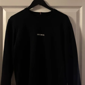 Svart tröja från Les Deux - Svart sweatshirt från Les Deux med enkel och clean design. Tröjan har rund halsringning, långa ärmar och vit broderad logga på bröstet. Perfekt för dig som gillar stilrena plagg med en modern vibe.