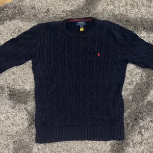 Mörkblå kabelstickad tröja Polo Ralph Lauren xs - Kabelstickad mörkblå tröja från Polo Ralph Lauren med klassisk rund hals och röd broderad logga på bröstet. Tröjan har långa ärmar och ribbade muddar vid ärmslut och nederkant. Perfekt för dig som gillar stilren och tidlös design. Storlek l i barnstorlek som är som xs eller xxs passar perfekt om du är 165-175