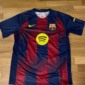 FC Barcelona hemmatröja 25/26 - Säljer en FC Barcelona matchtröja från Nike med klassiska blå och röda vertikala ränder. Tröjan har klubbmärke, Nike-logga, Spotify och UNHCR-tryck i gult samt Ambilight tv på ärmen. Tillverkad i lätt och ventilerande polyester.