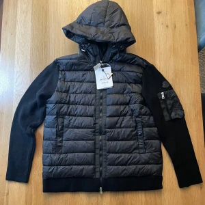 Svart Moncler pufferjacka med huva - Svart pufferjacka från Moncler med stickade ärmar och vadderad front. För mer info skriv jackan passar till dom som är 175-185 