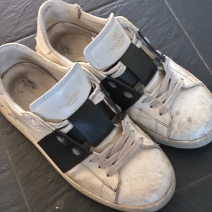 Valentino Garavani Open sneakers - Snygga Valentino Garavani Open sneakers i vitt skinn med bred svart läderdetalj över mitten. Klassisk rund tå, vita snören och tydlig logga på plösen. Sulan är mönstrad och märkt med Valentino. Perfekta för dig som gillar lyxiga och trendiga sneakers.