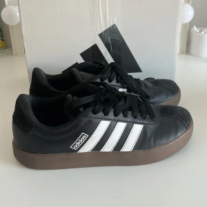 Adidas VL Court 3.0 sneakers svart/vit - Adidas VL Court 3.0 sneakers i svart med klassiska vita ränder på sidorna och brun platt sula, tillverkade i skinn och syntetmaterial. Använda i början av sommaren men slutade ganska snabbt, kan finnas diskreta defekter men annars som nya. 