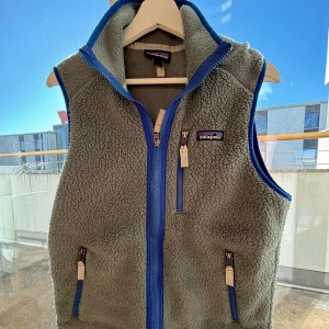  fleeceväst från Patagonia - Säljer en fleeceväst från Patagonia i storlek S. Västen har blå detaljer och dragkedjor framtill samt på fickorna. Den har en hög krage och ett mjukt, fluffigt material som är perfekt för lager-på-lager. Patagonia-loggan sitter på bröstet.