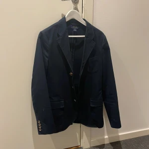Mörkblå kavaj från Polo Ralph Lauren - Säljer en stilren mörkblå kavaj från Polo Ralph Lauren. Den har en klassisk design med två knappar fram och fyra dekorativa knappar vid ärmsluten. Perfekt för både formella och semi-formella tillfällen. Kavajen är i mycket bra skick och passar perfekt för höst och vår. Storlek XL i junior vilket motsvarar ca S i herr!