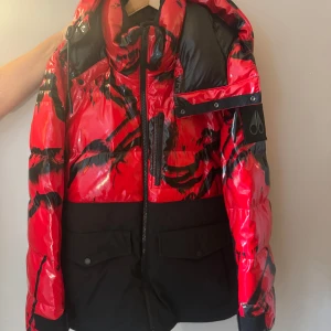 Röd och svart pufferjacka Moose Knuckles - Snygg pufferjacka från Moose Knuckles i rött och svart med coolt abstrakt mönster. Jackan har hög krage, huva, dragkedja framtill och flera fickor. Materialet är glansigt och jackan har en oversized vibe, perfekt för kalla dagar.