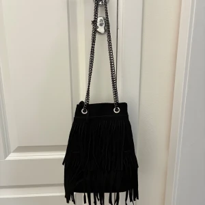 Svart mocka axelväska med fransar - Snygg svart axelväska i äkta mocka, tillverkad i Italien. Väskan har coola fransdetaljer och kedjeremmar i metall. Perfekt storlek för det viktigaste och stängs med magnetknapp. Insidan har ett fack med dragkedja. Använd en gång. 