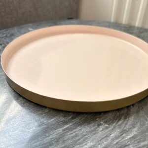 Bricka gör smink elr annat  - Söt rund bricka i ljusrosa med guldfärgad kant och botten. Brickan har en stilren design och är utrustad med halkskydd på undersidan. Passar utmärkt som serveringsbricka eller dekoration. Några få mindre ytliga repor, men annars i gott skick. 