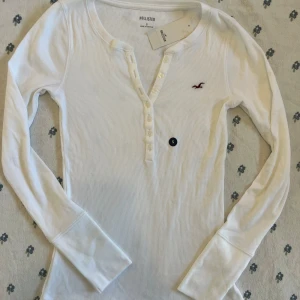 Vit ribbad långärmad Henley tröja Hollister - Supersnygg vit långärmad limited edition 2000s  vintage style henley tröja från Hollister med ribbad struktur och knappdetaljer framtill. Tröjan har en liten broderad logga på bröstet och är tillverkad i mjuk bomullsmix. Perfekt för lager-på-lager och chill vardagsstil. Lite genomskinlig bra att ha med linne under. 