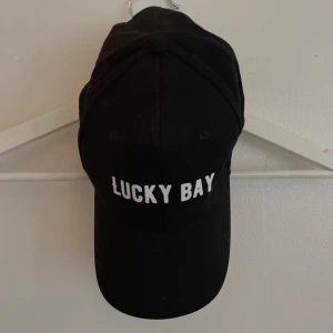 Svart keps Lucky Bay Black Hill - Svart keps från Black Hill med texten 'LUCKY BAY' i vitt framtill. Klassisk böjd skärm och justerbar rem baktill. Tillverkad i 100% bomull för en skön och luftig känsla. Perfekt accessoar för en cool och avslappnad stil.