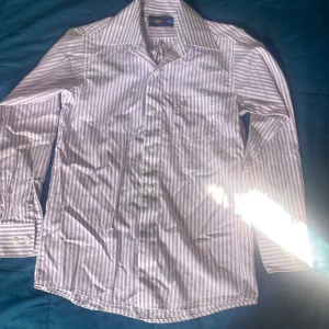 Randig skjorta från Manchester Junior - Snygg skjorta med lila och vita vertikala ränder från Manchester Junior. Klassisk krage, bröstficka med broderad logga och långa ärmar med dubbla knappar vid manschetten. Skjortan är i mjuk bomull och har en clean, fräsch look.