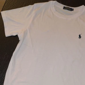 Vit t-shirt från Polo Ralph Lauren - Vit klassisk t-shirt från Polo Ralph Lauren med broderad marinblå logga på bröstet. T-shirten har rund halsringning och korta ärmar. Tillverkad i mjuk bomull för en skön känsla och clean stil. ( stryker t shirten inann frakt )