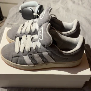 Grå Adidas Campus sneakers mocka - Snygga Adidas Campus sneakers i grå mocka med vita detaljer och klassiska tre ränder på sidan. Skorna har vit sula, breda vita skosnören och Adidas-logga på plösen. Perfekta för dig som gillar retrostil och streetwear.