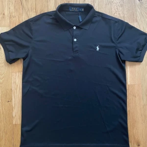 Svart pikétröja från Ralph Lauren - Klassisk svart pikétröja från Ralph Lauren med vit broderad logga på bröstet. Tröjan har krage, knäppning med två knappar och korta ärmar. Tillverkad i bomullspiké som ger en snygg och stilren look.