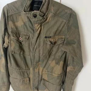 Camouflage vindjacka från Maison Scotch - Säljer en grön camouflage vindjacka från Maison Scotch. Jackan har flera fickor framtill med knappar, hög krage och dragkedja. Tillverkad i bomull och polyester, perfekt för dig som gillar en cool och avslappnad stil.