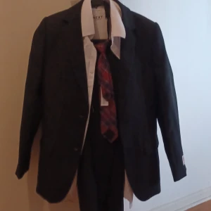 Svart barn kostymset från Opposuits & Next - Säljer ett stilrent svart barn kostymset med kavaj från Opposuits, matchande byxor och 2st  vit skjorta från Next. Till setet ingår även en röd och blå rutig slips. Kavajen har två knappar och klassiska fickor. Perfekt för dig som vill vara extra snygg på speciella tillfällen.Storlek 134-140.