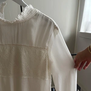 Vit plisserad klänning med spetsdetaljer - Superfin vit klänning med plisserade partier och breda spetsdetaljer över bröstet. Hög krage med volangkant och långa ärmar i tunt tyg. Perfekt för dig som gillar romantisk och elegant stil.