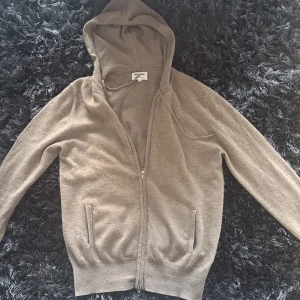 Beige zip-hoodie i kashmir från Soft Goat - Mysig beige hoodie från Soft Goat i lyxig kashmir. Tröjan har dragkedja framtill, huva med snörning och två fickor. Perfekt för dig som gillar en chill och stilren look. Långärmad och ribbade muddar för extra komfort.
