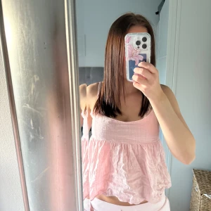 Ljusrosa volangig linnetopp och rosett där bak - Supersöt ljusrosa linnetopp med volangig nederdel och tunna axelband. Toppen har en luftig och flowy passform, perfekt för varma dagar. Materialet känns mjukt och lätt mot huden.