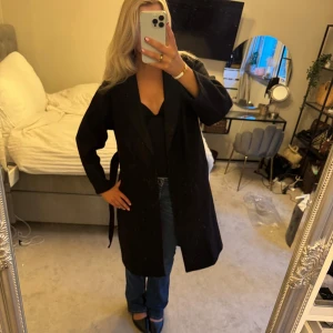 Svart kappa med bälte - Snygg svart kappa med bälte i midjan och klassisk krage. Långärmad och rak modell som ger en stilren look. Perfekt för dig som vill ha en tidlös och enkel jacka till höst och vinter.