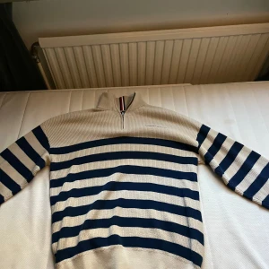 Randig stickad tröja med half zip - Beige och marinblå randig stickad tröja från Gant med half zip och hög krage. Tröjan har långa ärmar och ribbade muddar. Klassisk och stilren design med Gant-logga broderad på bröstet. Perfekt för lager-på-lager under kyliga dagar.