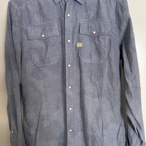 Blå mönstrad skjorta från G-Star RAW - Snygg blå skjorta från G-Star RAW med diskret mönster, två bröstfickor med knappar och klassisk krage. Skjortan har vita knappar framtill och på ärmsluten. Perfekt för dig som gillar stilrena detaljer och streetwear-vibe.