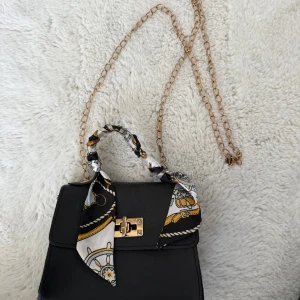 Svart handväska med kedja och scarf - Stilren svart handväska med guldfärgad kedja som axelrem och snyggt mönstrad scarf knuten runt handtaget. Väskan har ett guldfärgat lås framtill och är tillverkad i ett slätt material. Perfekt accessoar för att lyfta din outfit.