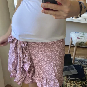 Rosa blommig omlottkjol med volang - Supersöt kort omlottkjol i ljusrosa med små vita blommor. Kjolen har volangkant och knyts i sidan för en extra gullig look. Perfekt för varma dagar och ger en riktigt somrig vibe. Tunn och lätt i materialet, passar perfekt till sneakers eller sandaler.