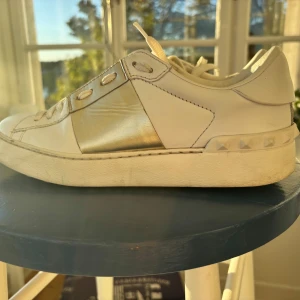 Valentino Garavani sneakers med gulddetalj - Snygga vita sneakers från Valentino Garavani med bred guldfärgad rem över vristen och coola nitar på sulan. Skorna har snörning, rund tå och är tillverkade i skinn. Perfekta för dig som vill sticka ut med exklusiva detaljer och lyxig känsla. Priset kan diskuteras 