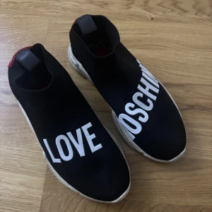 Love Moschino svarta sneakers med hjärta - Svarta sneakers från Love Moschino med vit text på ovandelen och röda hjärtan bak på hälen. Skorna har en chunky vit sula och är tillverkade i stickad syntet för en sock-liknande passform. Perfekta för dig som vill sticka ut med coola detaljer.