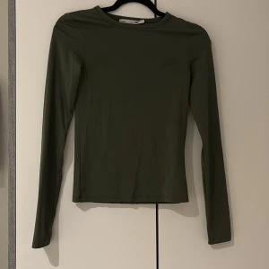 Mörkgrön långärmad tröja 157 - En stilren mörkgrön långärmad tröja från 157. Tröjan har en slim passform och är tillverkad i ett mjukt, stretchigt material som känns skönt mot huden. Perfekt att matcha med jeans eller kjol för en clean look.