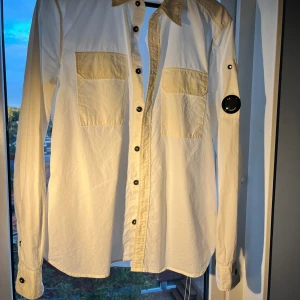 Vit overshirt från C.P. Company - Vit overshirt från C.P. Company med svarta knappar, två stora bröstfickor och den ikoniska linsen på vänster ärm. Skjortan har klassisk krage och detaljer i en något ljusare beige ton på axlar och fickor. Perfekt för dig som gillar streetwear med stil.