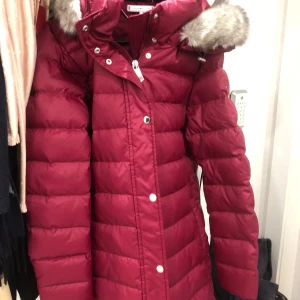 Tommy Hilfiger-jacka (mörkröd) i storlek M - Mörkröd jacka från Tommy Hilfiger med avtagbar huva och fuskpäls. Jackan har dragkedja och knappar framtill, samt praktiska fickor. Den har klassiska Hilfiger-detaljer på insidan och är oanvänd. Galge är inte inkluderad. Troligen säljs inte längre.
