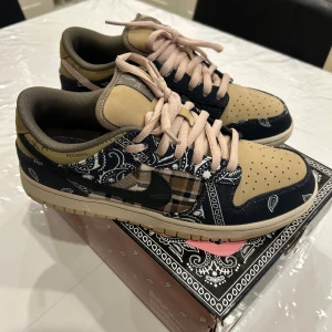 Nike SB Dunk Low Travis Scott - Nike SB Dunk Low Travis Scott med beige mocka, mörkblå och rutiga paneler samt bandanamönster. Skorna har rosa snören, svart och rosa swoosh samt platt sula. Unik design med paisley och rutigt tyg, perfekt för dig som vill sticka ut. Skorna är i mycket bra skick knappt använda. Skriv gärna på minsta fundering, frågor eller extra detaljbilder/videor!