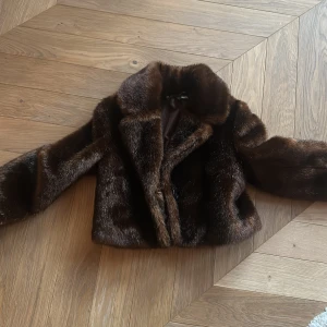 Mörkbrun pälsjacka - Säljer en snygg mörkbrun pälsjacka med klassisk krage och knäppning framtill. Jackan har en lyxig, glansig finish och är fodrad på insidan. Den har en fin croppad längd som passar perfekt till t.ex ett spetslinne! 