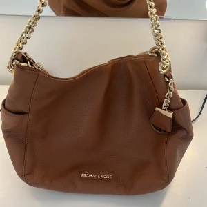 Brun axelväska Michael Kors i skinn - Snygg brun axelväska från Michael Kors i äkta skinn med guldfärgade kedjedetaljer och dragkedja. Väskan har flera fack, logga framtill och ett klassiskt hänglås med MK-emblem. Perfekt storlek för allt du behöver ha med dig.