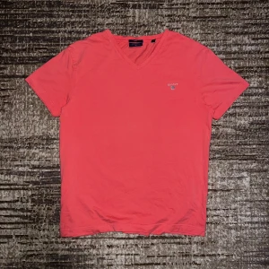 Korallröd GANT t-shirt med v-ringning - Snygg korallröd t-shirt från GANT med v-ringad hals och broderad logga på bröstet. Modellen är fitted och har korta ärmar. Tillverkad i mjuk bomull som känns skön mot huden. Perfekt för dig som gillar stilrena och klassiska plagg.