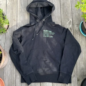 Helmut Lang hoodie - Svart hoodie från Helmut Lang med broderad text på bröstet. Klassisk känguruficka, snörning i huvan och ribbade muddar. Perfekt för en chill och stilren look.