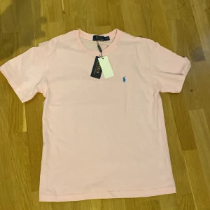 Ljusrosa t-shirt från Polo Ralph Lauren - Snygg ljusrosa t-shirt från Polo Ralph Lauren med klassisk blå logga broderad på bröstet. T-shirten har rund halsringning och korta ärmar. Tillverkad i mjuk bomull för en skön känsla. Perfekt för en clean och stilren look.