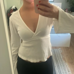 Vit omlottopp med knytband - Snygg vit topp med långa ärmar och v-ringning. Toppen har knytband bak i ryggen som knyts baktill och lätt rynkad struktur i tyget. Perfekt för en stilren och fräsch look.