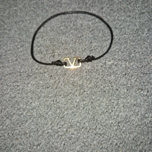 Valentino armband med svart snöre - Stilrent armband från Valentino med svart justerbart snöre och en liten guldfärgad logga i metall. Perfekt accessoar för att lyfta din outfit med en touch av lyx och enkelhet.