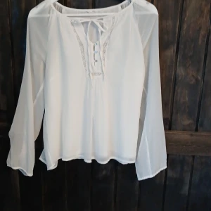Vit blus med spets och knyt - Säljer en luftig vit blus med transparenta långärmar, dekorativa knappar framtill och fina spetsdetaljer längs halsringningen. Blusen har knytband vid halsen och är tillverkad i ett tunt, flowy material som ger en romantisk vibe.
