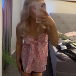 Rosa blommig topp med rosett - Supersöt topp i rosa och vitt med blommigt mönster och stor rosett framtill. Axelbandslös modell som ger en fräsch och somrig vibe. Perfekt för dig som vill sticka ut med en feminin och lekfull stil.