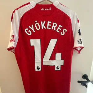 Gyökeres Arsenal Hemmaställ 25/26 - Röd och vit Arsenal fotbollströja från Adidas med Gyökeres och nummer 14 på ryggen. Tröjan har korta ärmar, broderat klubbmärke, Premier League-tryck och detaljerade mönster i tyget. Tillverkad i ventilerande funktionsmaterial.