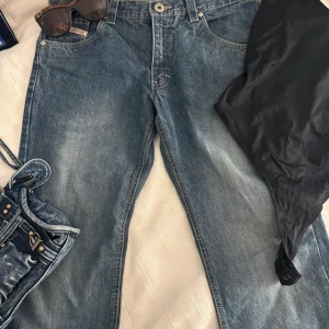 Blå jeans från Denim Traveller - Säljer ett par blå jeans från Denim Traveller med snyggt slitna detaljer och kontrastsömmar. Bakfickorna har broderade mönster i gult. Jeansen har rak passform och vintage look. 