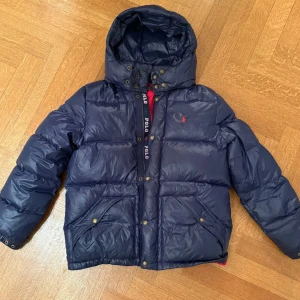 Mörkblå pufferjacka från Polo Ralph Lauren - Mörkblå pufferjacka från Polo Ralph Lauren med röd insida och broderad logga på bröstet. Jackan har huva, stora fickor med tryckknappar och dragkedja framtill. Klassisk och sportig look med Polo-detaljer på insidan.