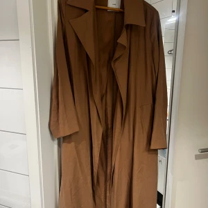 Brun trenchcoat från Mango - Stilren brun trenchcoat från Mango med klassisk krage och långa ärmar. Jackan har en avslappnad passform och är tillverkad i ett mjukt material som känns skönt mot huden. Perfekt för lager-på-lager och snygg till både jeans och sneakers.