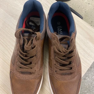 Bruna sneakers från Muzzo, storlek 42 - Snygga bruna sneakers från Muzzo i storlek 42. Skorna har en mix av mocka och syntet med perforerade detaljer på tån och sidorna. De har bruna snören, vit sula och mörkblå foder på insidan. Perfekta för en stilren och avslappnad look.