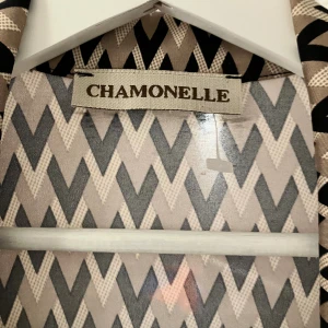 Klänning från Chamonelle - Snygg klänning från Chamonelle med coolt zigzag-mönster i beige och svart. Klänningen är gjord i ett mjukt, glansigt material som känns skönt mot huden. Perfekt för dig som gillar att sticka ut med grafiska prints.