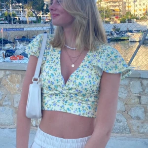 Blommig croppad topp - Topp med ljusgul botten och små blommor i lila, blått och grönt. V-ringad omlottfront med volangiga korta ärmar.